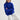 Woman wearing a blue sweater and white pants sitting on a stool against a white background