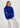 Woman wearing a blue sweater and white pants sitting on a stool against a white background