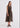 A woman modeling a sleeveless midi dress with a sheer, patterned black mesh fabric featuring vertical stripes in muted red, brown, and beige tones, paired with black strappy flat sandals against a plain white studio background.