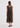A woman viewed from behind wearing a sleeveless midi dress with a sheer mesh overlay and a vertical geometric pattern in olive green, black, and rust red. She is wearing black strappy sandals against a plain white studio background.