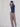 A model stands against a draped white fabric backdrop, wearing a black long-sleeve top, black beanie, tan cropped trousers, and black sandals.