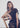 A woman with shoulder-length brown hair sits on a wooden chair against a white draped fabric backdrop, wearing a dark brown one-shoulder top and matching dark bottoms.