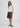 A woman modeling a sleeveless cream knit top with lace detailing at the collar and buttons, paired with brown shorts and brown strappy sandals, against a plain light gray studio background.